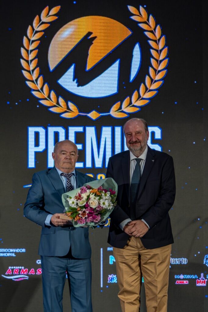 Two men in suits on a stage at an awards ceremony; one presents a bouquet to the other, with a large 'PREMI' logo and laurel wreath behind them.
