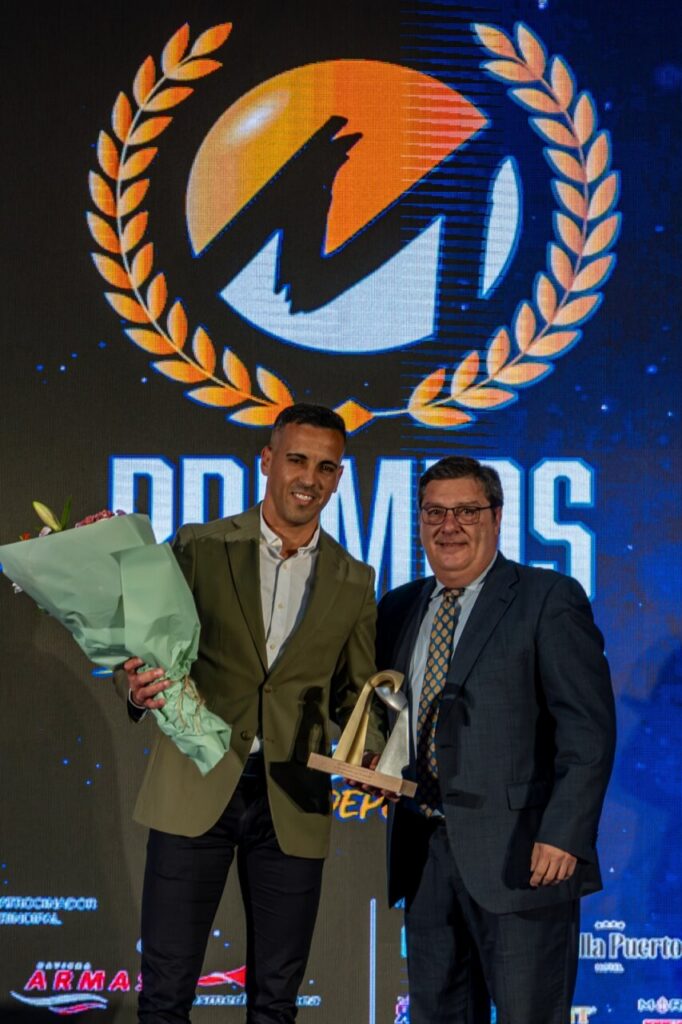 Two men in suits on a stage, one presenting a bouquet and a trophy; they smile for the photo with a large award logo and 'AWARDS' backdrop behind them.