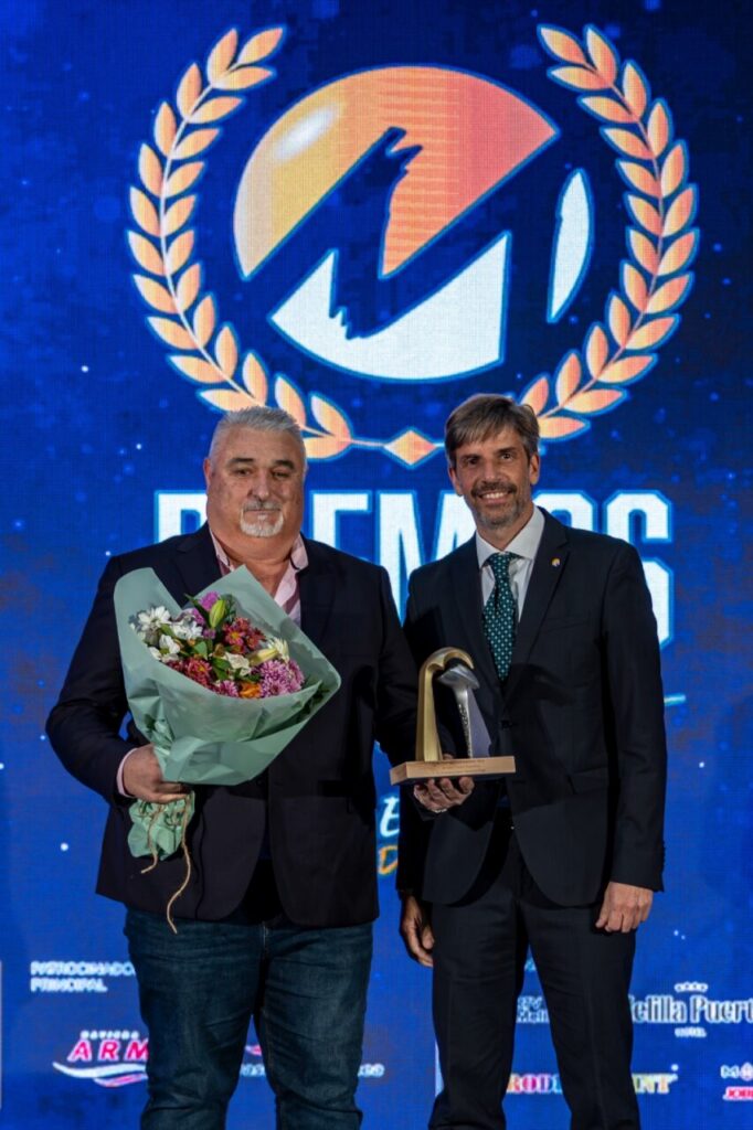 Dos hombres sosteniendo un trofeo y un ramo de flores en un evento de premiación