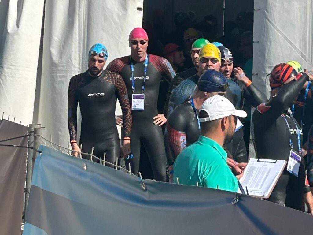 Group of swimmers in wetsuits and colorful swim caps stand at the race start, an official with a clipboard in the foreground observing them.