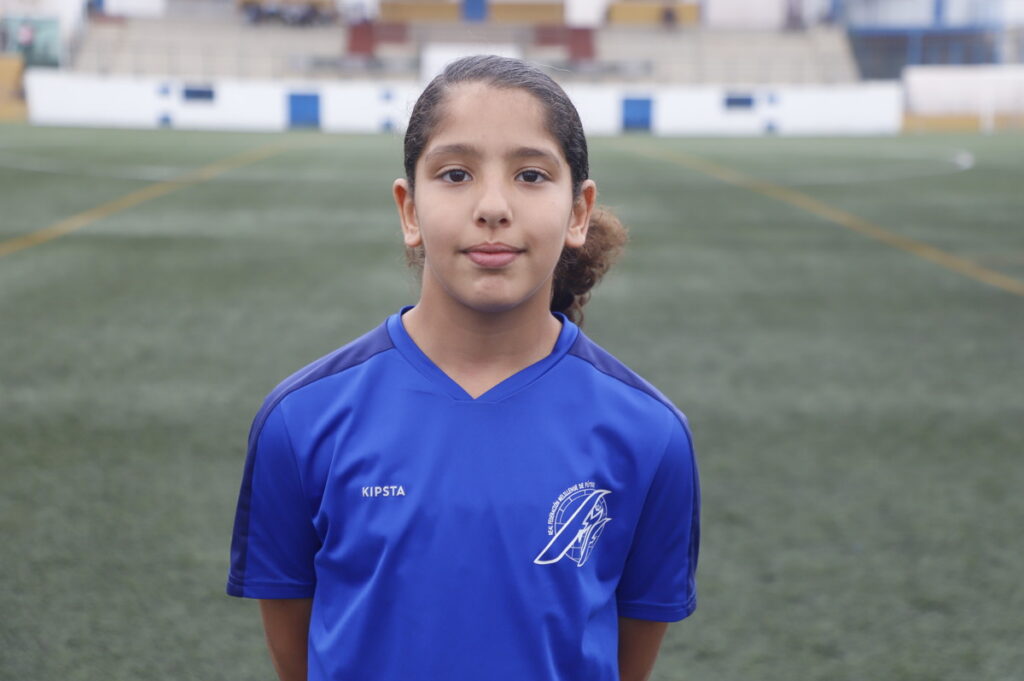 Young girl in a blue jersey standing on a soccer field, facing the camera with a neutral expression.