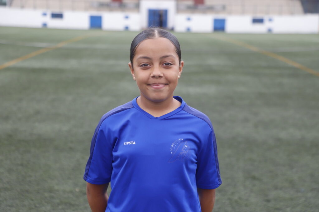 Girl in a blue sports jersey standing on a football/soccer field looking at the camera.