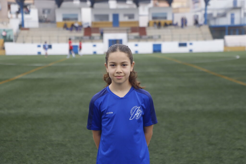 Young girl in a blue soccer jersey standing with hands behind her on a soccer field, stadium seating in the background.