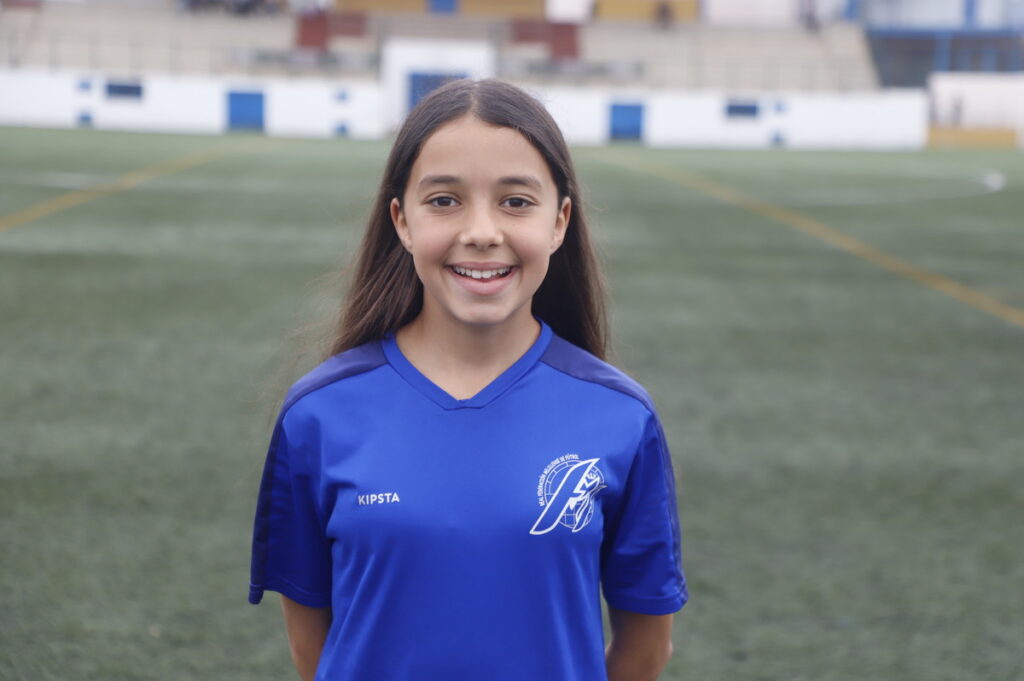 Smiling girl in a blue Kipsta soccer jersey on a grassy field, facing the camera.