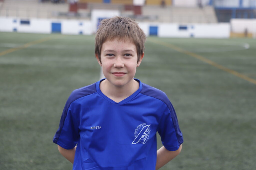Young boy in a blue soccer jersey standing on a grassy field with stadium stands behind him, hands clasped behind his back.