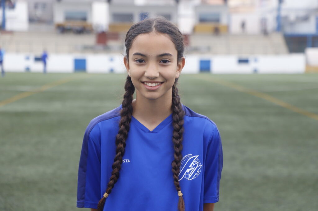 Smiling girl in a blue soccer jersey with two braids on a field background.