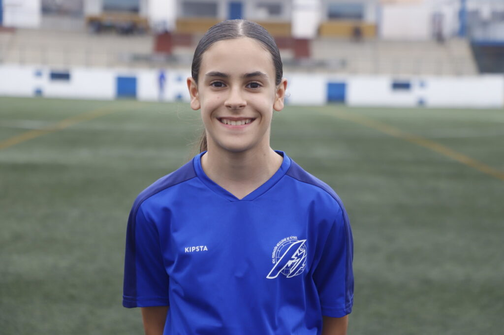 Young girl in a blue sports jersey standing on a soccer field, smiling at the camera.
