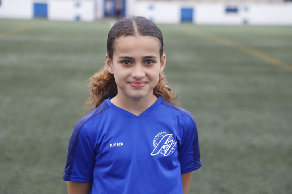Young girl in a blue soccer jersey standing on a grassy field, facing the camera with a slight smile.