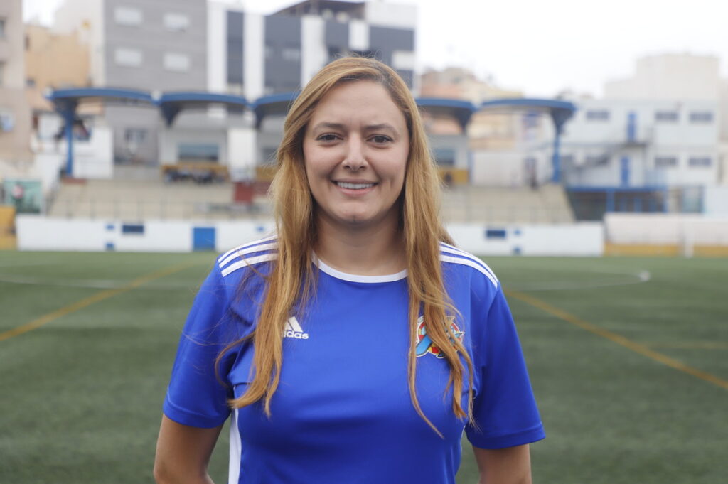 Smiling woman in a blue soccer jersey stands on an empty stadium field with bleachers in the background.