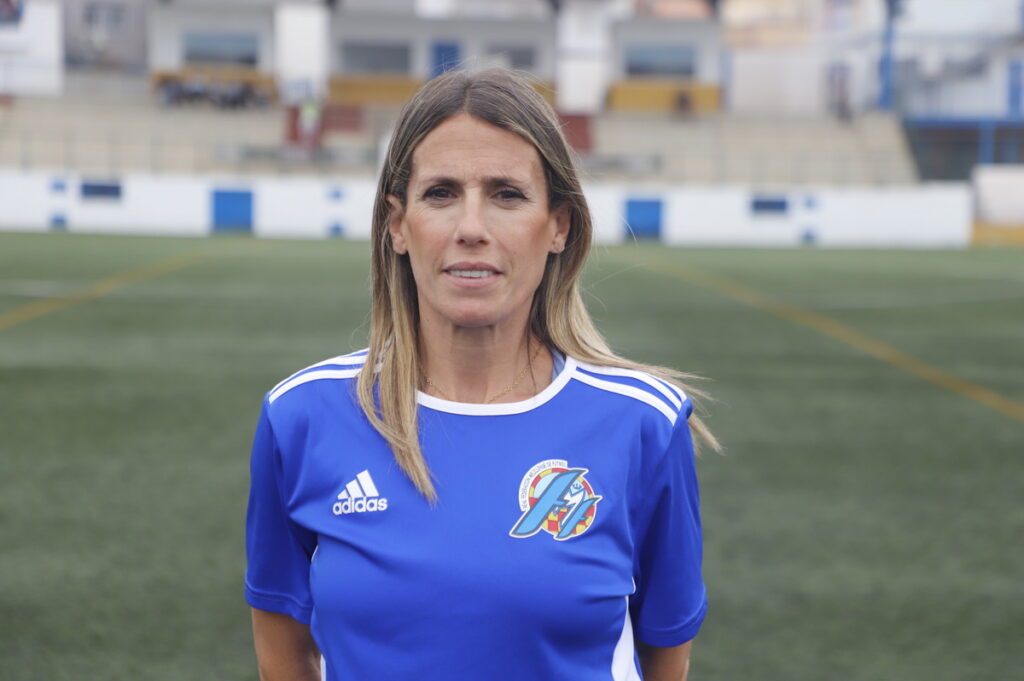 Portrait of a woman soccer player in a blue Adidas jersey with team crest, standing on a green turf field with bleachers in the background