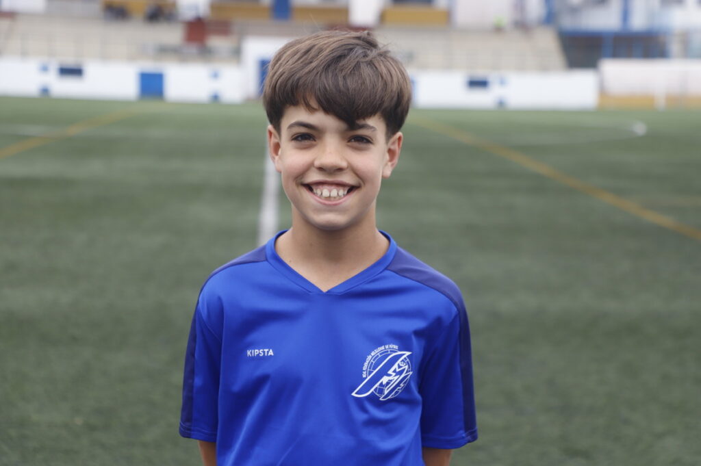 Smiling boy in a blue soccer jersey standing on a grassy field with stadium seating in the background simulating a youth match.