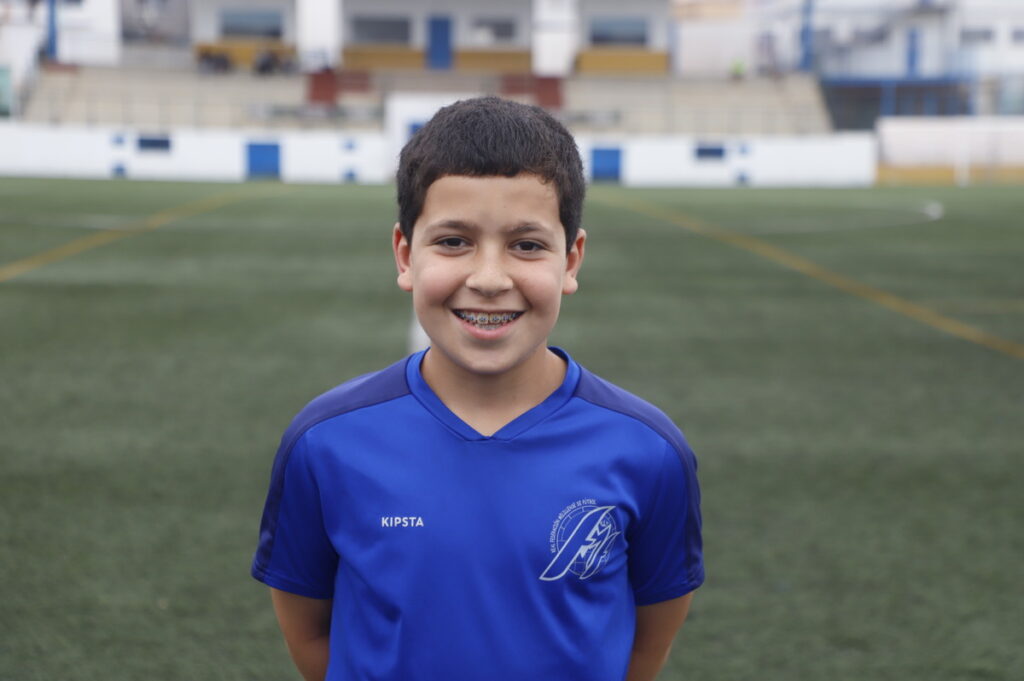 Smiling boy with braces in a blue soccer jersey on a field, with blurred stadium stands behind him.