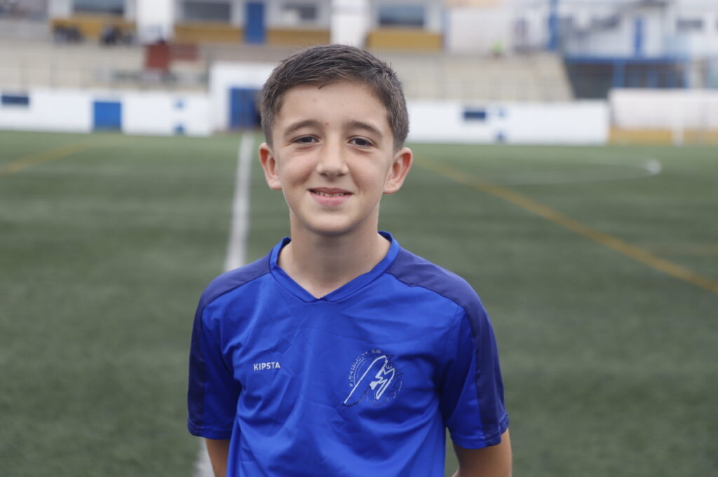 Portrait of a smiling boy in a blue soccer jersey standing on a grassy field near stadium seats.