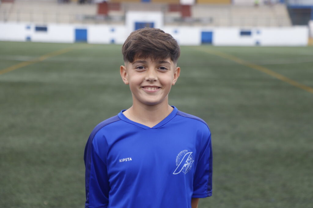 Smiling boy in a blue soccer jersey standing on a green artificial turf field with stadium seating in the background.