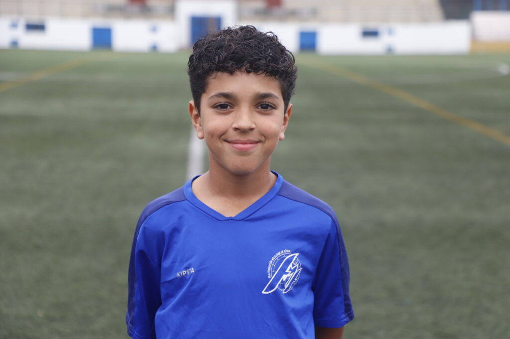 Smiling boy in a blue soccer jersey standing on a green field with goal lines in the background