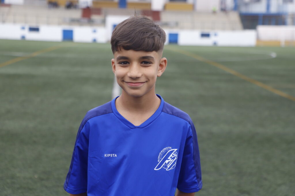 Smiling boy in a blue soccer jersey standing on a soccer field with blurred stadium seating behind him.