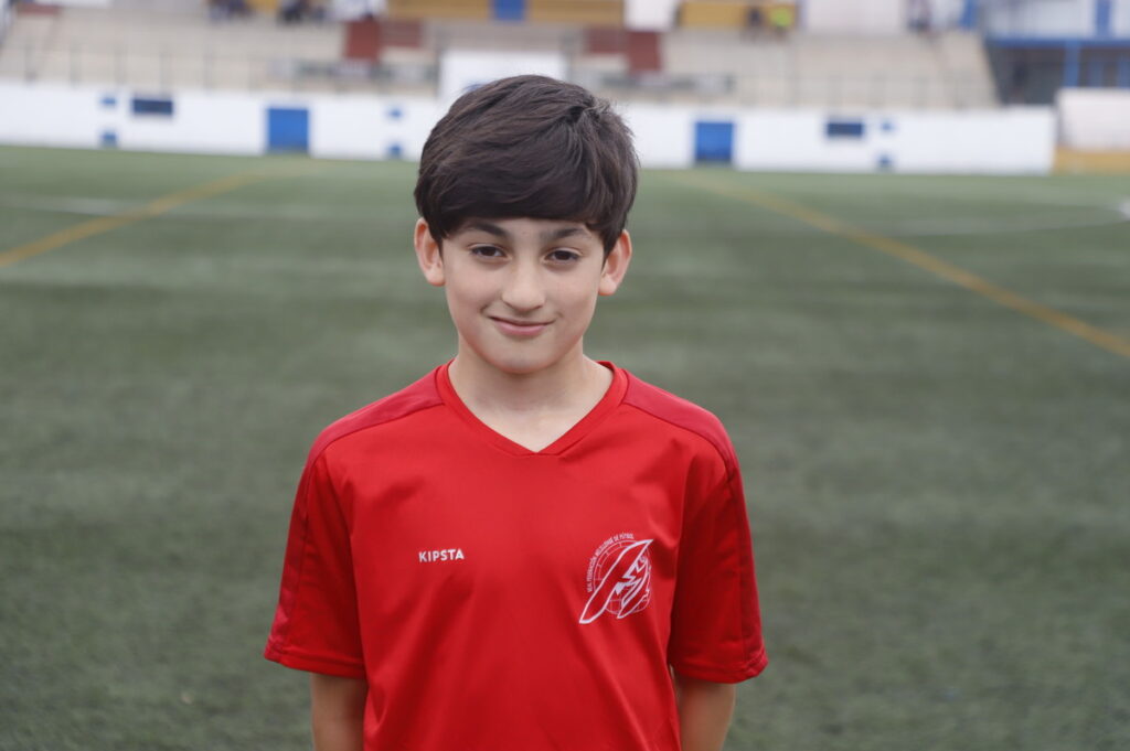 Young boy in a red soccer jersey standing on a green football field.