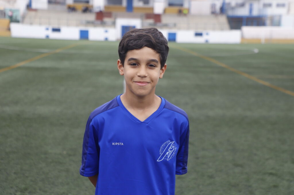 Young boy in a blue soccer jersey standing on an outdoor field, smiling at the camera, with stadium seating in the blurred background.