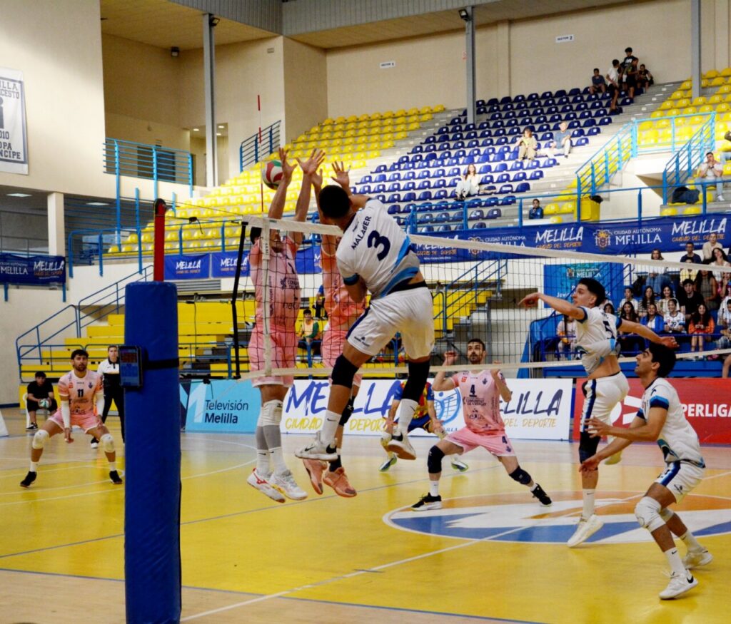 Volleyball players jump at the net during a match in a yellow-blue arena, with a pink team blocking and a white-jersey attacker in mid-air.