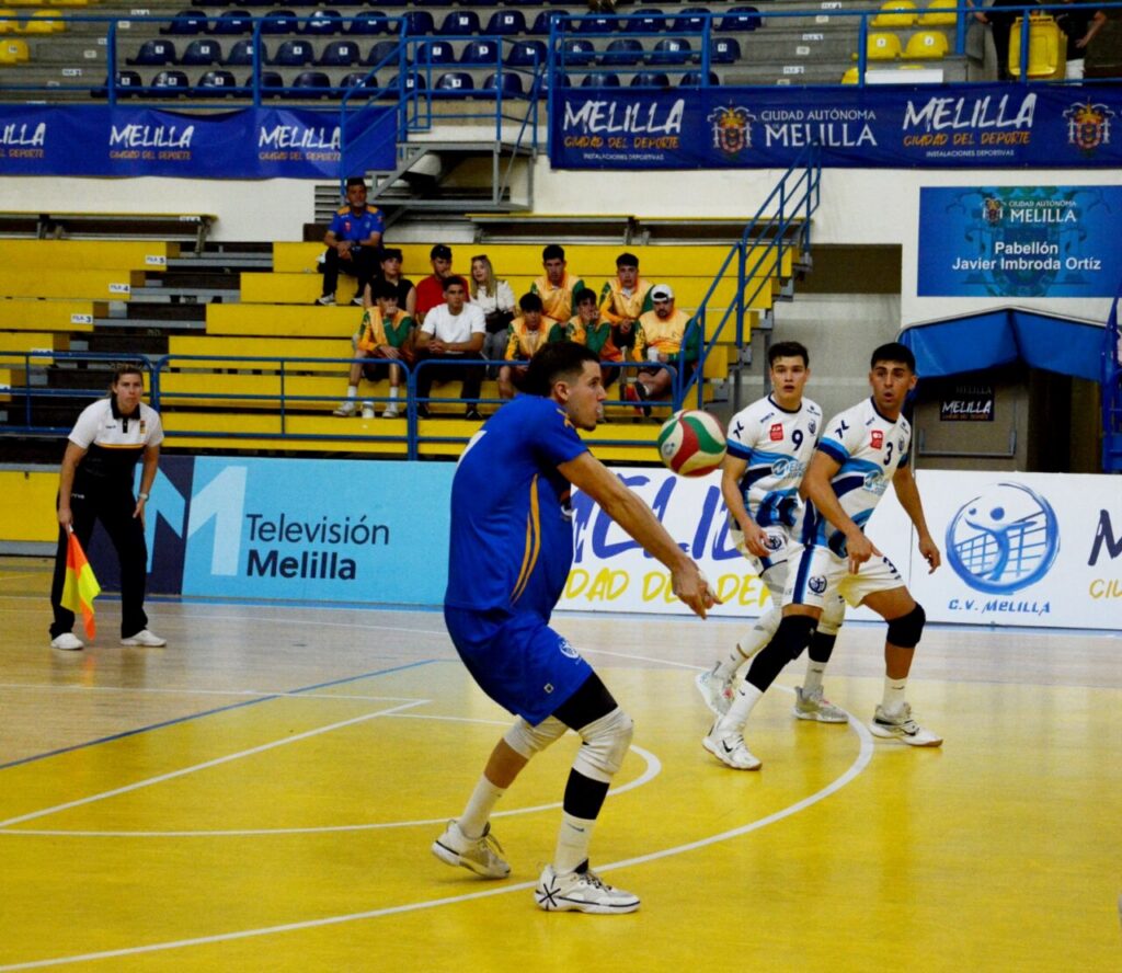 Futsal match on a yellow indoor court with blue and white players chasing the ball as spectators watch from yellow bleachers.