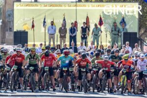 Group of cyclists at the starting line in colorful jerseys, with spectators and officials on a stage behind them.