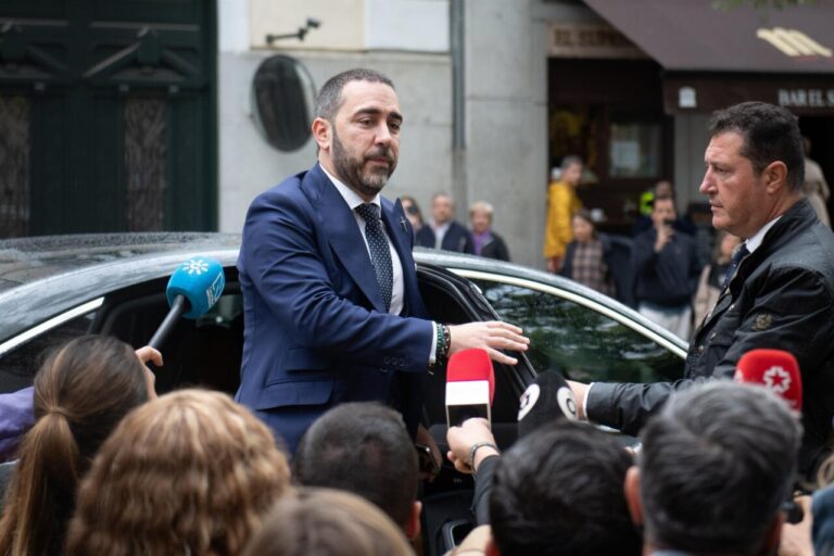 Man in a navy suit steps out of a black car as reporters with colorful microphones surround him on a city street.