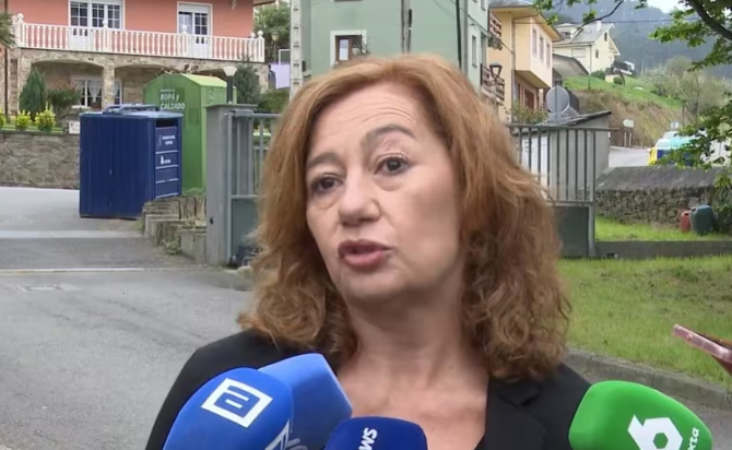 Woman with curly red hair speaks to reporters on a street, microphones from multiple outlets in the foreground, colorful houses in the background.