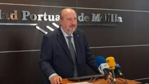 Older man in a dark suit at a podium speaking to reporters, with blue and yellow microphones in front.