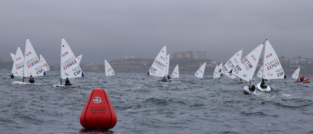 Group of small sailboats racing around a red buoy on a choppy sea under a gray, overcast sky near a coastal city skyline with distant buildings.