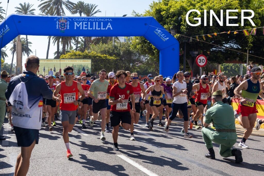 Runners surge at the Melilla city marathon start under a blue inflatable arch, a photographer kneels to capture the moment.