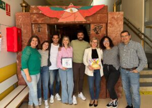 Eight adults posing together in a hallway in front of a brown stone art structure featuring an eye with wings, all smiling at the camera.