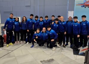 Group of young athletes in matching blue jackets posing for a team photo indoors, with luggage visible nearby.