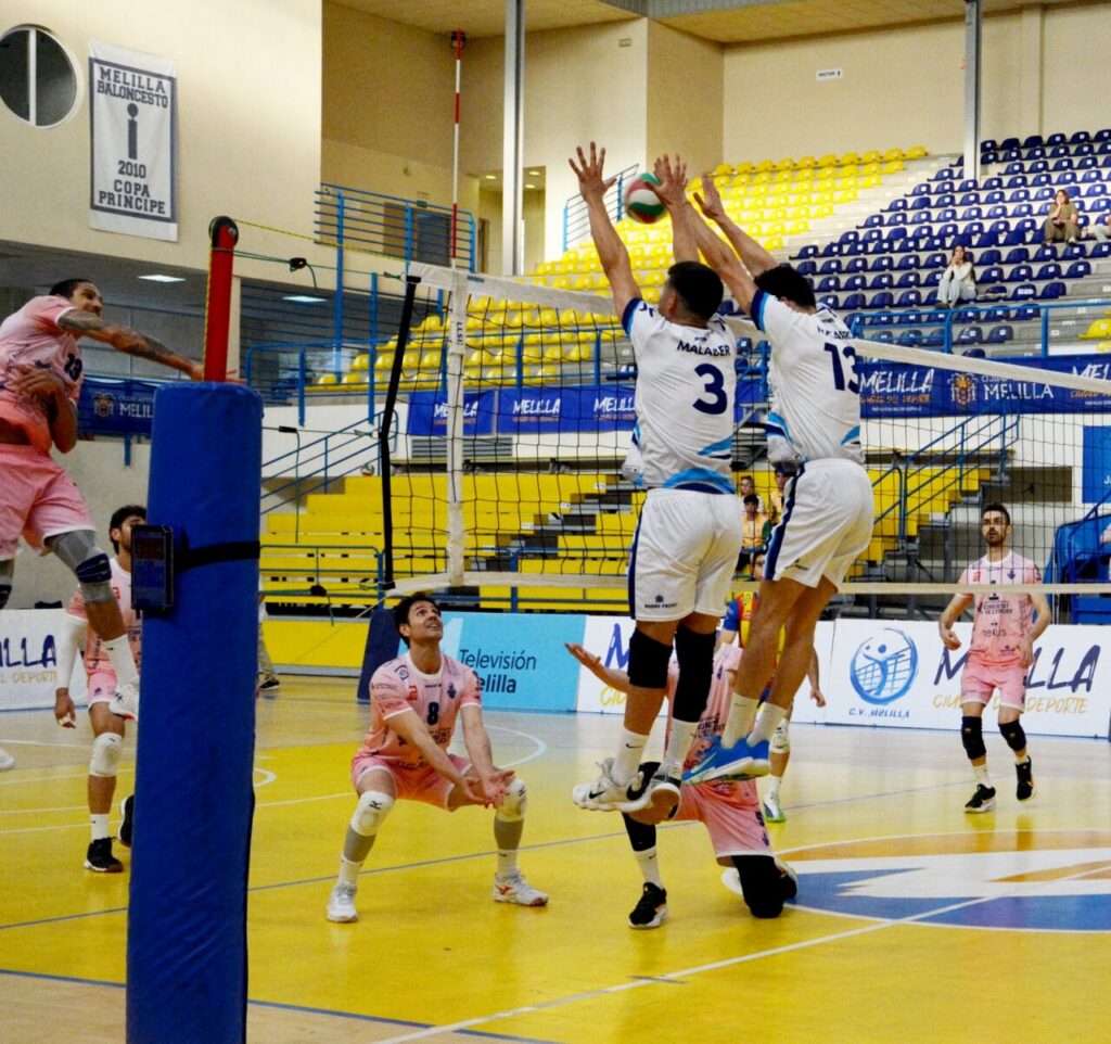Volleyball players at the net jumping to block a ball during a match in a colorful indoor arena with yellow and blue seats in the background.