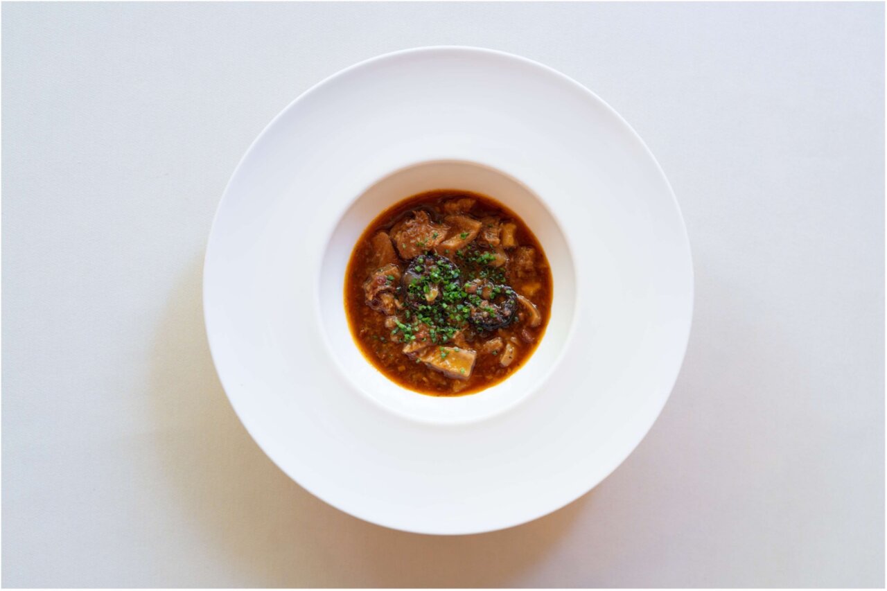 Top-down view of a white bowl with brown beef stew and parsley on a white plate