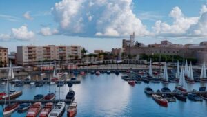 Marina with many small sailboats and colorful boats moored in calm blue water, harbor buildings in the background under a cloudy sky