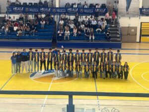 Group of basketball players in yellow and blue jerseys posing for a team photo on a gymnasium court with spectators in the stands behind them and banners above.
