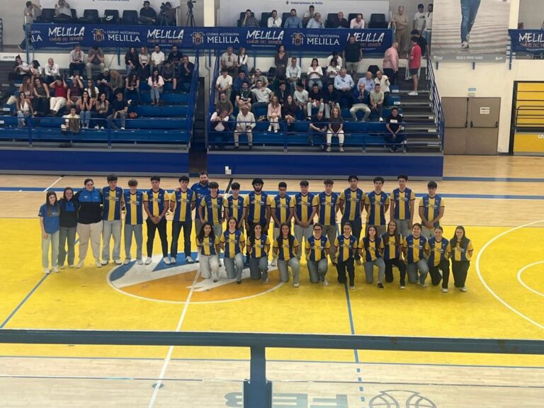 Group of basketball players in yellow and blue jerseys posing for a team photo on a gymnasium court with spectators in the stands behind them and banners above.