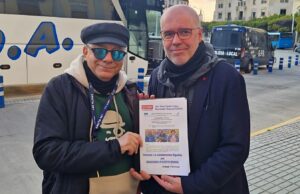 Two men stand together outdoors, one holding a document or flyer facing the camera near buses and a street scene.