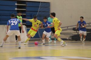 Futsal match with players in yellow and blue jerseys contesting a pink ball near the goal on an indoor court inside a gym.