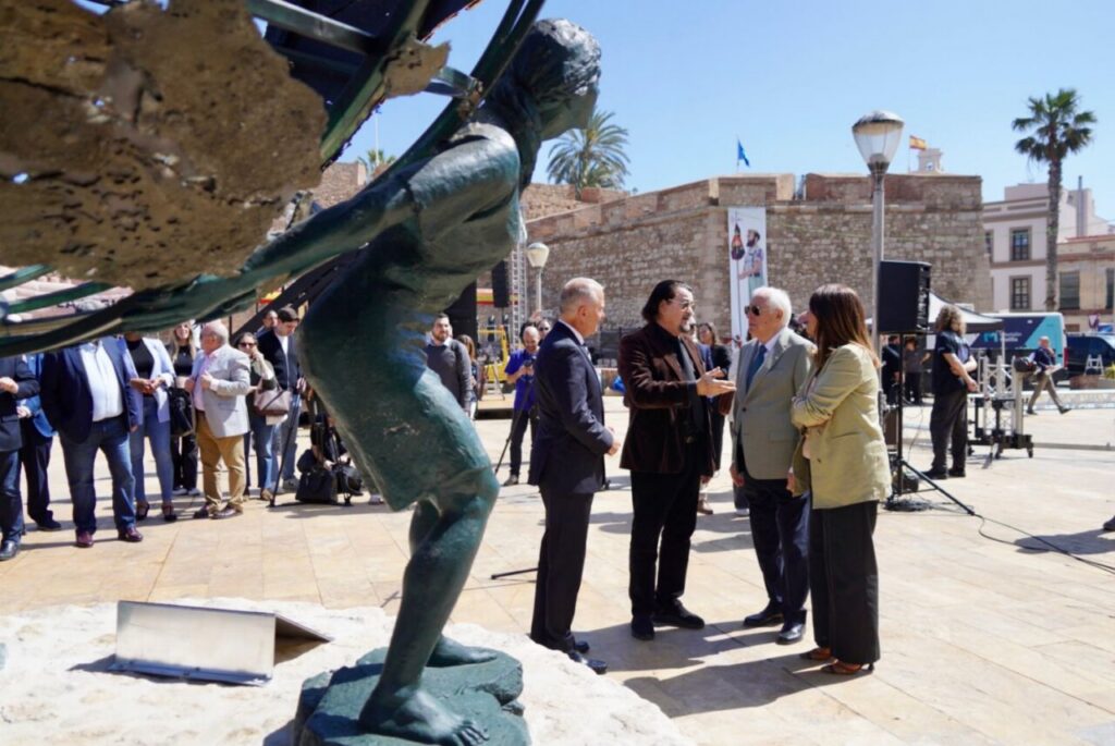 Formal group of people in conversation near a bronze statue in an open plaza with stone walls and palm trees behind them.