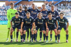 Soccer team posing for a group photo on the field in navy uniforms with a neon-clad goalkeeper, stadium seats in the background.