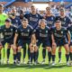 Soccer team posing for a group photo on the field in navy uniforms with a neon-clad goalkeeper, stadium seats in the background.