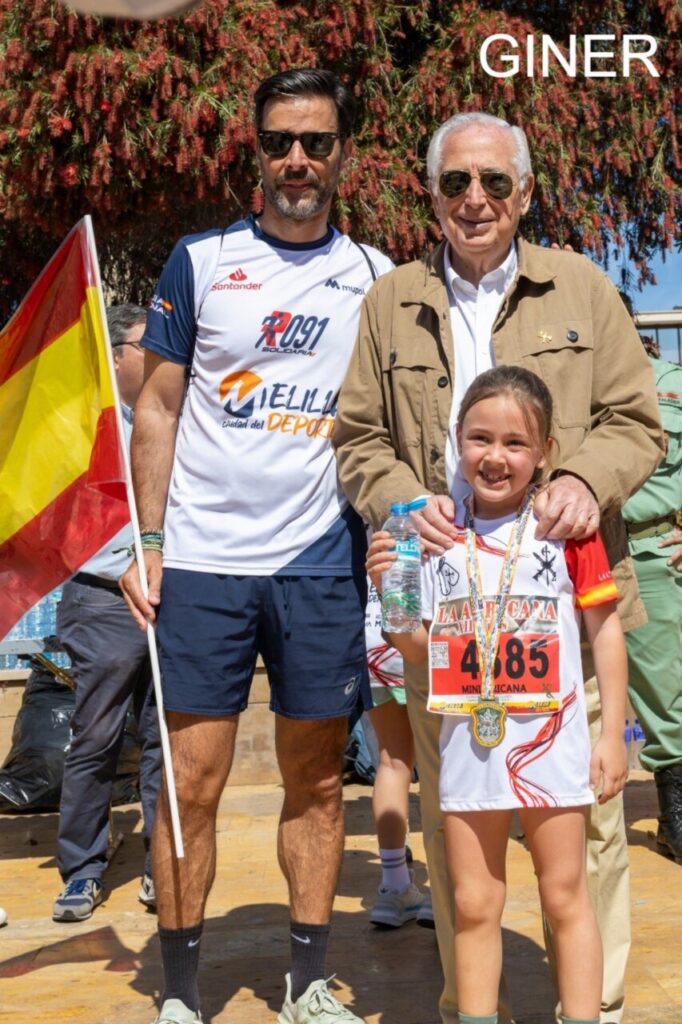 Young girl with race bib 4685 and a medal, holding a water bottle, posing with two men at a running event.