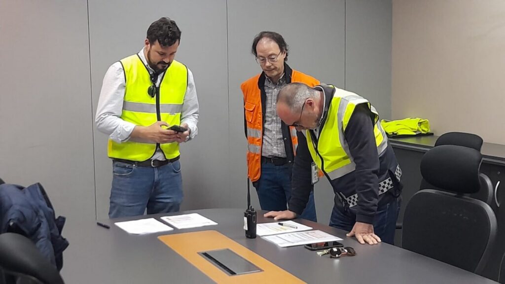 Tres hombres con chalecos reflectantes revisan documentos en una mesa