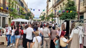 Crowded street market with stalls, shoppers, and colorful bunting overhead viewed along a sunny daytime street.