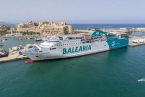 Large white Balearia passenger ferry docked in a harbor, turquoise water around, with a historic fortress and buildings in the background.
