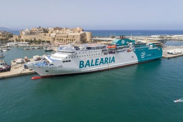 Large white Balearia passenger ferry docked in a harbor, turquoise water around, with a historic fortress and buildings in the background.