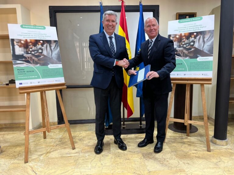 Two men in business suits shake hands in a formal setting with flags, posters, and certificates nearby, marking a signing or partnership event.