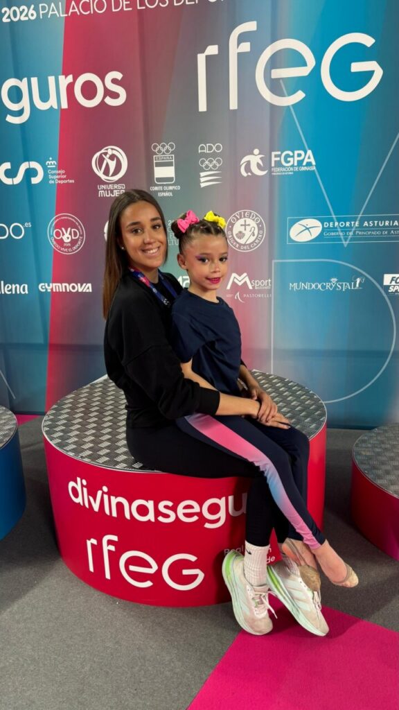 Two young girls sit closely on a red circular platform at a gymnastics event, smiling with colorful banners behind them.