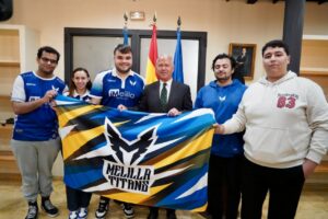 Six people stand indoors holding a colorful Melilla Titans banner, with two flags and a portrait in the background.